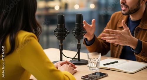 Two people recording podcast in modern studio. Professional microphones capture animated conversation. Woman in yellow sweater listens while bearded man gestures expressively.