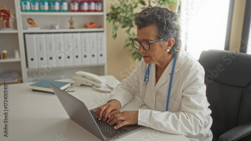 Wallpaper Mural Senior woman doctor with grey hair working on a laptop in a clinic office, surrounded by medical equipment and files. Torontodigital.ca