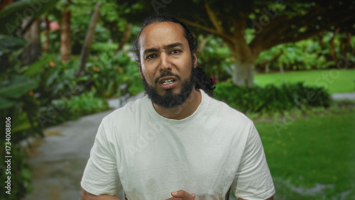 Man with hands clasped and worried face speaking on a park path in forest with green foliage and lawn; concern sincerity.
