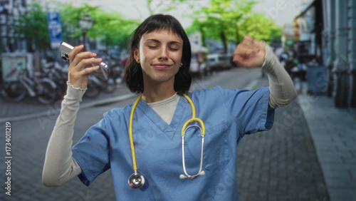 Woman nurse wearing blue scrubs and stethoscope raises phone and clenches fist in city street; pride celebration.
