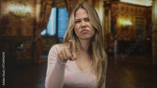 Young blonde woman pointing finger at camera in ornate hotel lobby with a stern expression and direct gaze; reprimand tension.