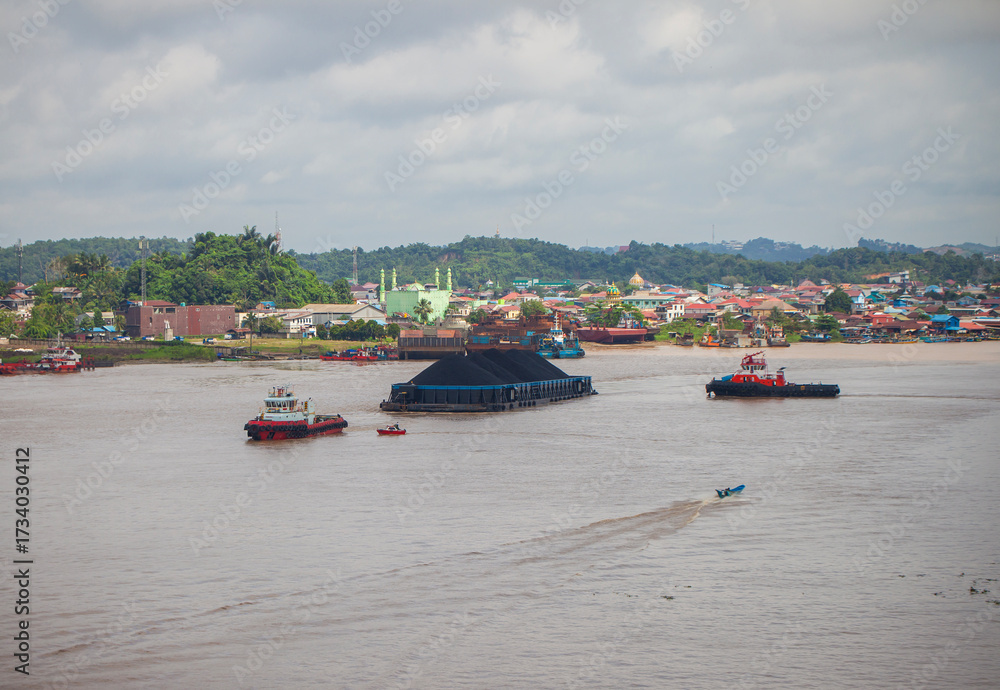Obraz premium Contrasting activity on the Mahakam River in Samarinda, East Borneo, Indonesia. A massive coal barge, guided by tugboats, passes a small local motorboat, showcasing the river's diverse traffic
