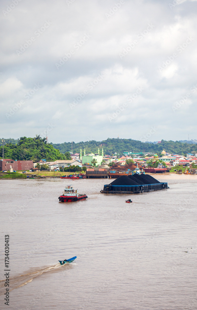 Obraz premium Contrasting activity on the Mahakam River in Samarinda, East Borneo, Indonesia. A massive coal barge, guided by tugboats, passes a small local motorboat, showcasing the river's diverse traffic