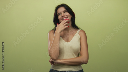 Photography Young hispanic woman in beige tanktop touching chin in pensive pose against green studio backdrop; contemplation