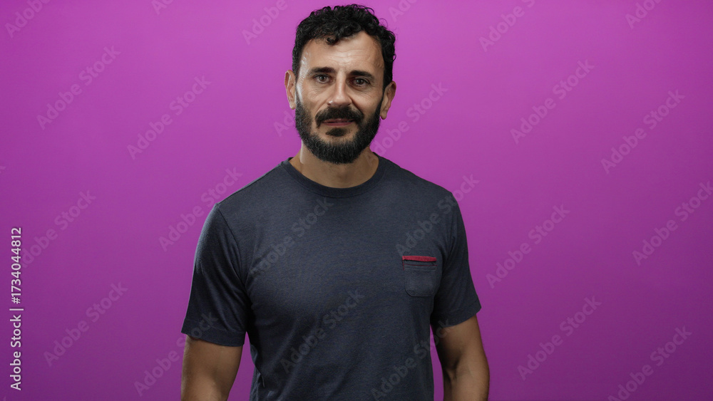 Naklejka premium Man standing against a vibrant pink background, wearing a casual grey shirt, exuding confidence and calmness, his dark beard and hair complementing the bright setting.