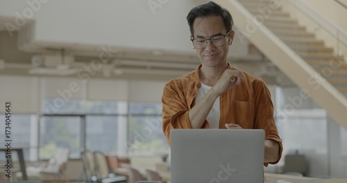 Senior Asian businessman wearing headset celebrates success during online meeting in modern co-working space. Remote work and business communication concept