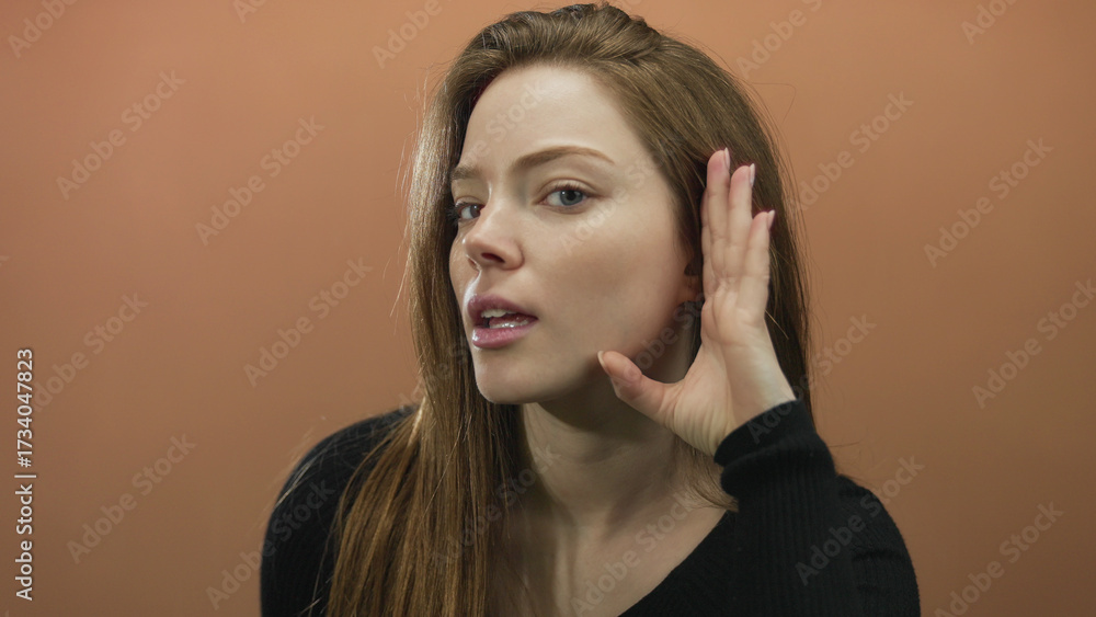 Fototapeta premium Woman with brown hair and serious expression cupping hand to ear against orange wall in studio; focused listening.
