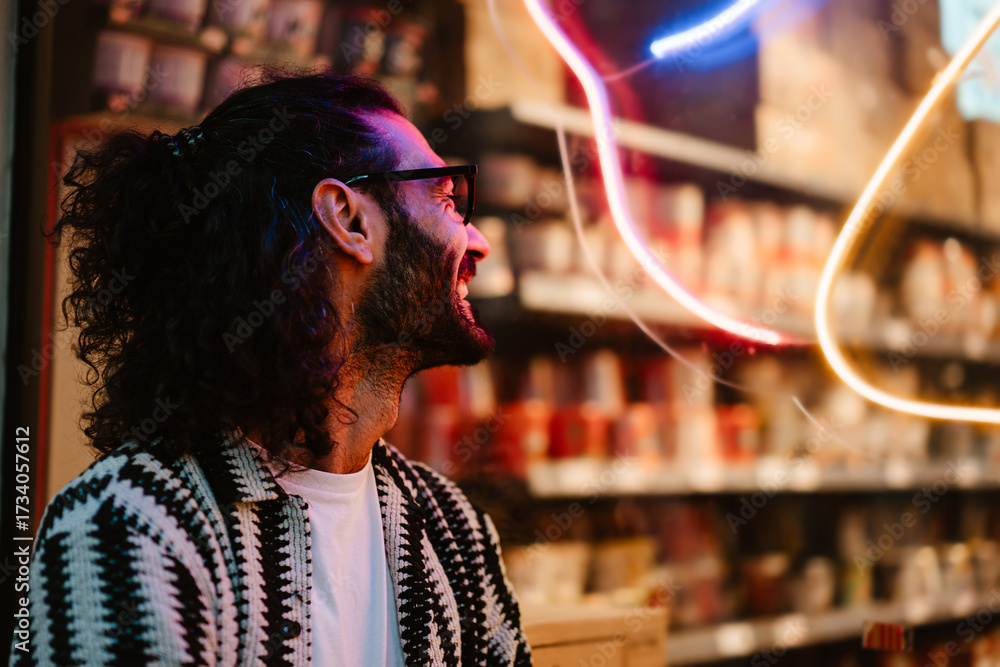 Obraz premium Man smiles looking away from neon window, surrounded by glowing lights, reflecting quiet joy and stylish tech lifestyle in night urban setting.