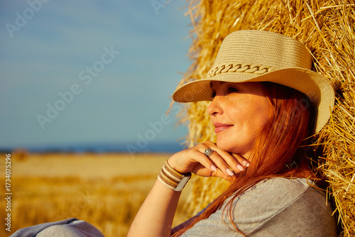 red-haired beautiful woman with hat in field with haystacks against blue sky