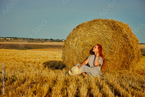 red-haired beautiful woman with hat in field with haystacks against blue sky