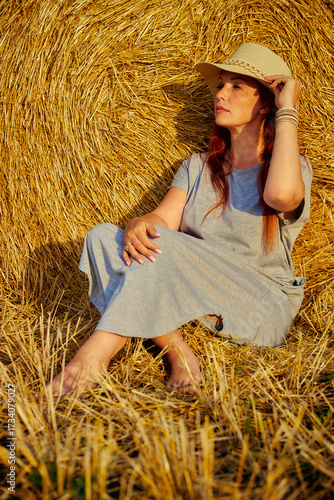 red-haired beautiful woman with hat in field with haystacks against blue sky