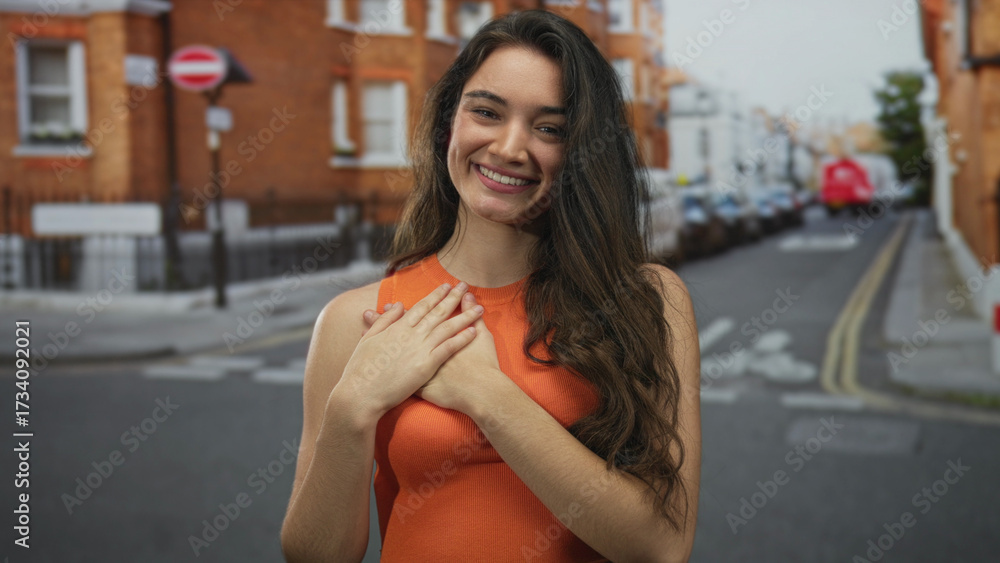 Fototapeta premium Hispanic woman wearing an orange sleeveless top clutching her chest on a narrow city street lined with brick buildings; gratitude.