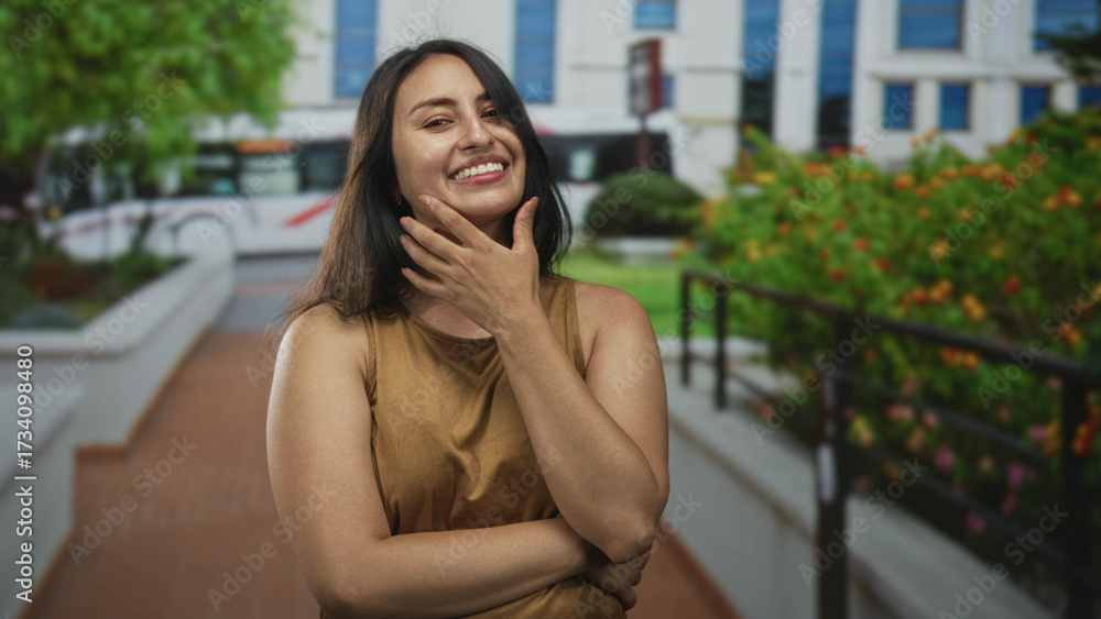 Fototapeta premium Young hispanic woman bites nails with hand to mouth on a building walkway near a bus and shrubs; playful confidence.