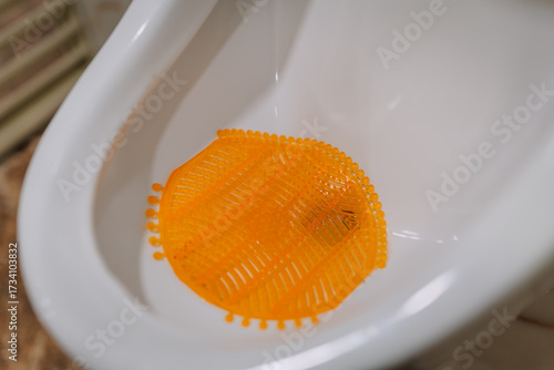 Photos Close-up of a white urinal with an orange urinal screen inside, typically used for hygiene and odor control in public or commercial restrooms