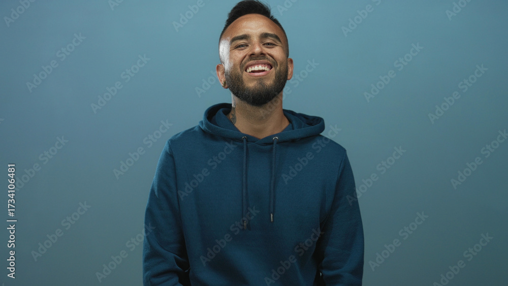 Fototapeta premium Man smiling showing teeth and beard in teal studio wearing blue hoodie, relaxed pose; joy warmth openness.