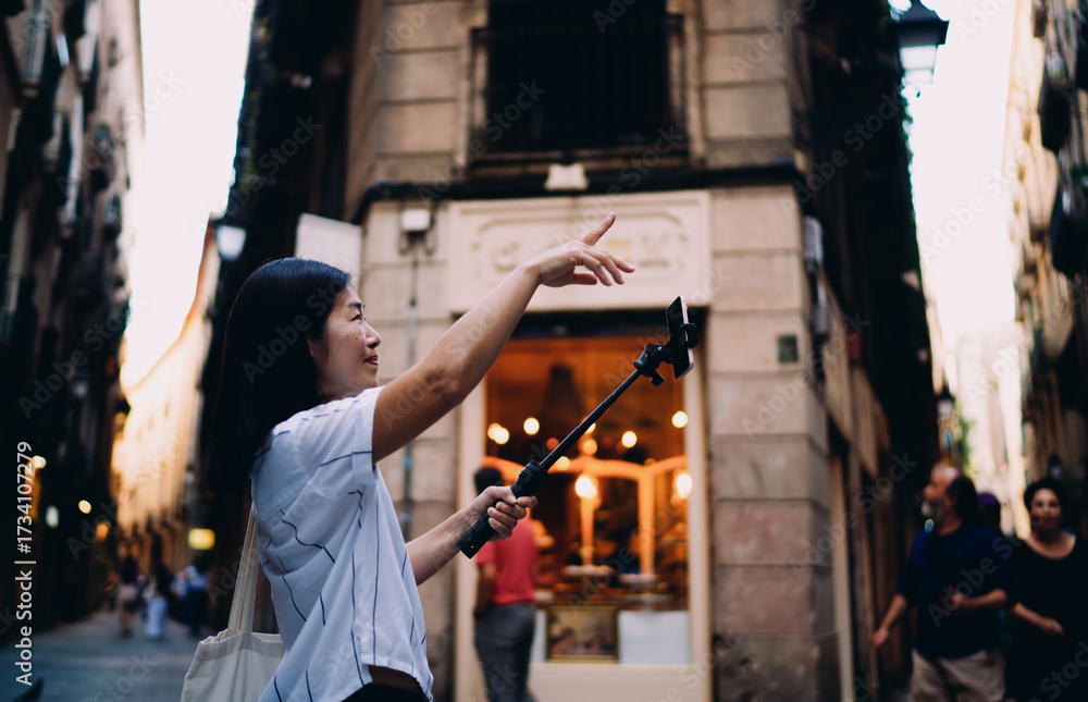 Obraz premium Asian woman with long dark hair talks to camera on smartphone stabilizer while walking down street, creating engaging vertical video content for social media or vlog audience.