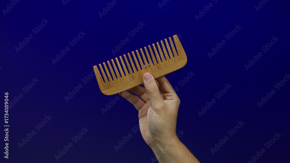Naklejka premium Man holding wooden comb against blue background, showcasing eco-friendly grooming product with caucasian male hand in focus, highlighting sustainability and personal care.