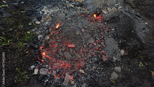 Smoldering red embers glow amid dark ash and stones — raw, intense, and earthy, evoking the quiet power of fire’s aftermath in a rugged, natural setting.