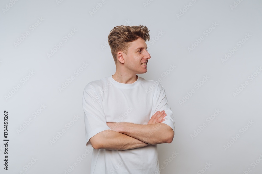 Obraz premium Young man stands with arms crossed, smiling confidently against a plain white background in a casual setting