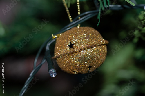 A glittering, gold-colored Christmas bell ornament, hanging among the green foliage of a festive holiday tree