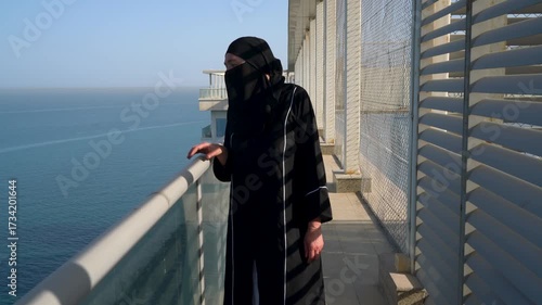 A woman dressed in traditional Arab clothes stands on the balcony with an amazing view of the coastline. She looks thoughtfully at the calm sea under the transparent sky, covering the moment.