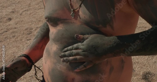 Young boy rubbing therapeutic blue clay mud on his stomach and arms for a natural skin care treatment while enjoying a sunny day on a sandy beach, promoting wellness and alternative therapy.