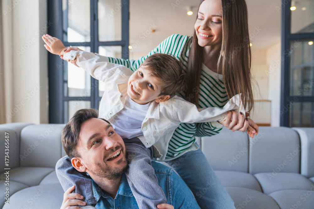 Obraz premium Happy laughing kid boy having fun with his family, sitting on father's shoulders at home. Portrait of young couple looking cheerful to each others sitting on sofa and spending time together with son