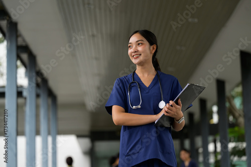 Asian doctor woman smiling, holding clipboard in hospital hallway