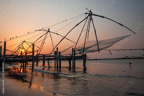 Carta da parati March 25, 2016, in Kochi, India, the iconic Chinese fishing nets rise against a golden sunset, their reflections on the water echoing Kerala’s timeless tradition, culture, and coastal heritage
