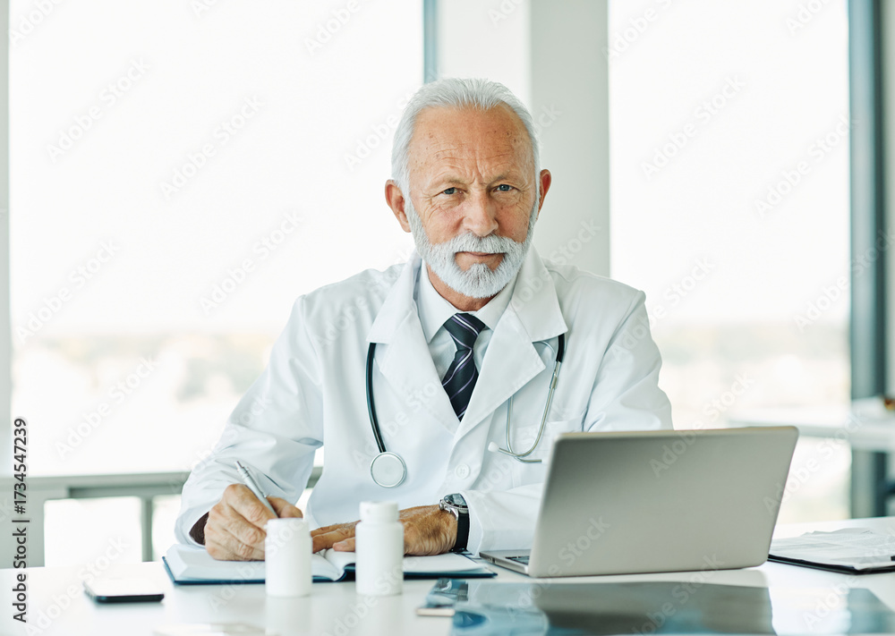 Obraz premium Portrait of a happy elderly male senior doctor using a laptop in his office in a hospital