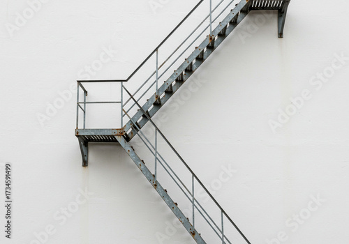 Durable industrial metal fire escape stairs ascend against a stark white textured building facade, suggesting urban grit and architectural detail.