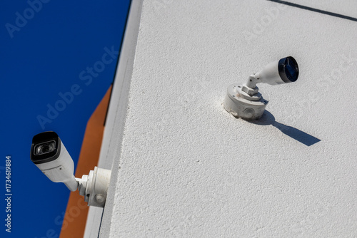 Two security cameras mounted on a building corner monitor the area, capturing footage against a bright blue sky