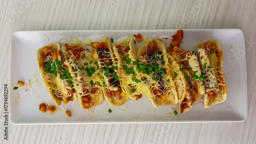 A delicious platter of mini tacos filled with savory ingredients, topped with melted cheese and fresh chives, served on a white plate on a wooden table, perfect for a party or a flavorful meal.
