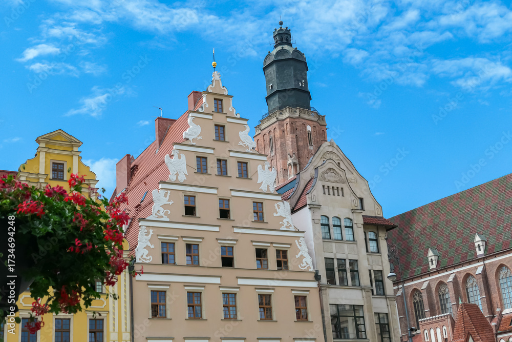 Fototapeta premium Ornate gabled buildings and St. Elizabeth’s Church dominate Wroclaw’s city center, with a lamppost and vibrant flowers in the foreground enhancing the picturesque urban landscape.