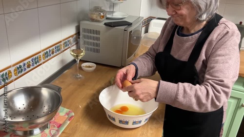 Senior woman with white hair cracking eggs to prepare Spanish omelette in rustic kitchen