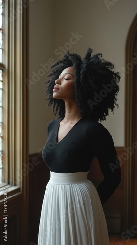 A woman with dark hair stands near a window, facing the light with her eyes closed. She wears a black top and a white skirt. She appears to be relaxing or meditating