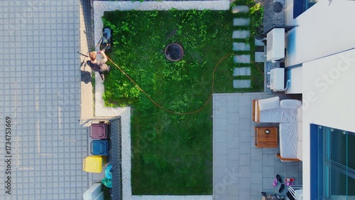 Man mowing green grass with electric lawn mower in backyard of modern house, top view. Person working in yard of suburban house. Concept of gardening, yard care and home maintenance