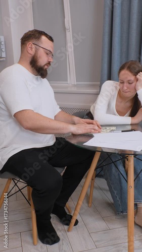 Focused young couple sitting at kitchen table, carefully managing household finances, meticulously counting dollars and collaboratively discussing budget strategies vertical video