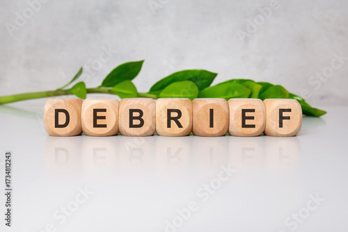 Wooden cubes spelling DEBRIEF on glossy white desk with green leaves in blurred background