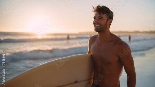 Handsome surfer smiles while holding surfboard at sunset on the beach