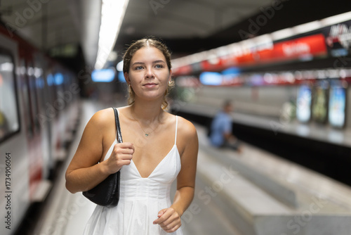 Positive young woman wearing white dress looking for convenient train in subway station