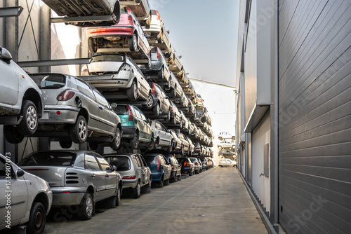 Stacked cars in scrapyard with multiple vehicles lined up on shelves. The scene shows rows of cars awaiting recycling or dismantling on sunny day