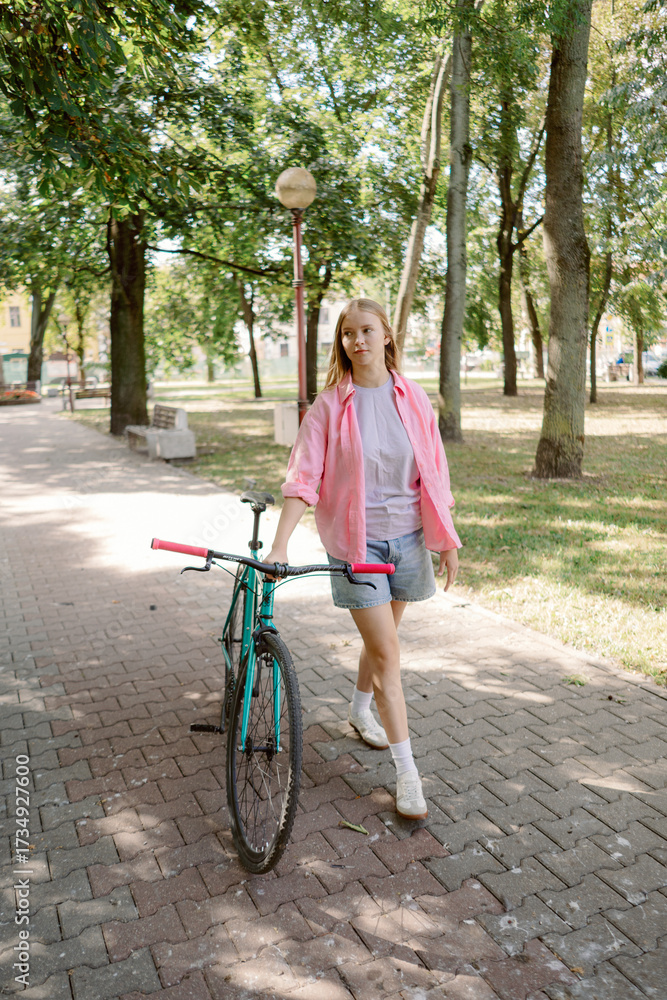 Naklejka premium A girl in a pink jacket and denim shorts strolls down a park path. She pulls her bright bike beside her as she enjoys the sunshine and greenery around her.