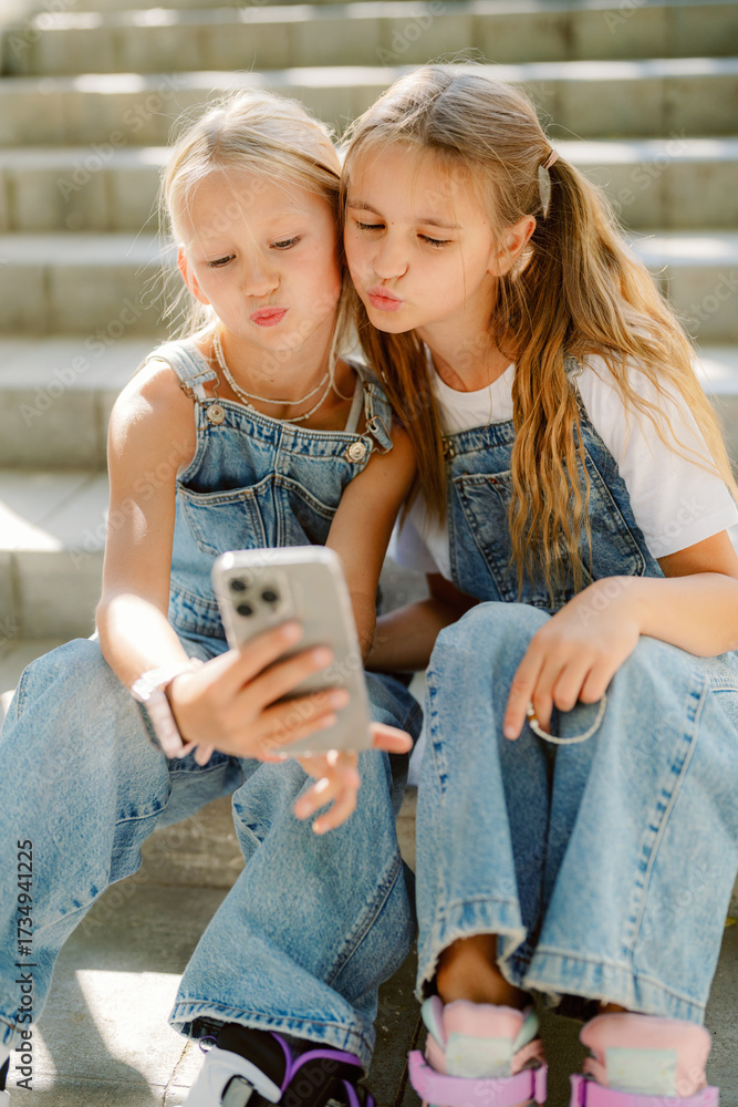 Fototapeta premium Two girls are sitting on stone steps, wearing denim overalls and making funny faces at their phone while capturing selfies. They are enjoying a joyful moment together.