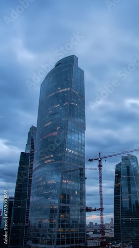 Modern glass skyscrapers and a crane against a dramatic, cloudy dusk sky in a sprawling city