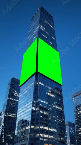 A towering glass skyscraper at dusk with a large, blank green screen billboard for advertising