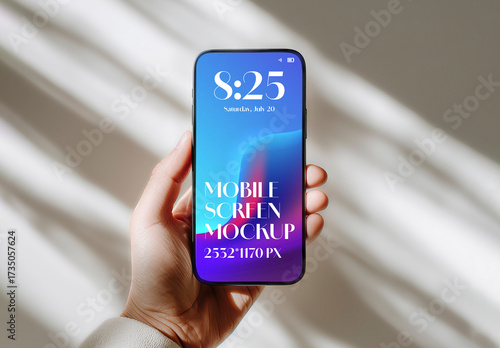 Hand holding a modern smartphone with a blank screen for a mockup against a wall with window shadows