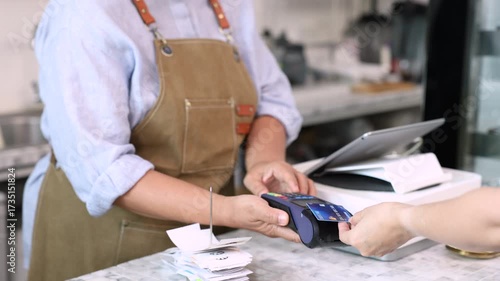 Senior entrepreneur cafe owner barista holds POS terminal while customer swipes creditcard, Asian retired female in apron smiling at counter, demonstrating cashless payment inside modern coffee shop