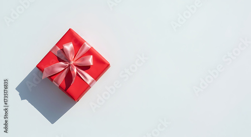 Minimalist top view of a single red gift box with a pink satin bow on a white background, with hard sunlight shadow and ample copy space for holiday, birthday, or valentine's day.