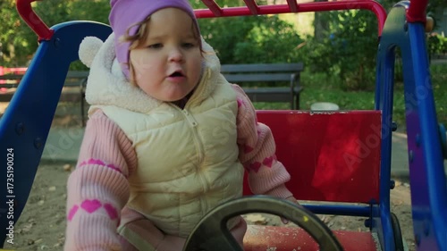 Child plays on a red playground toy in a sunny park surrounded by trees and benches
